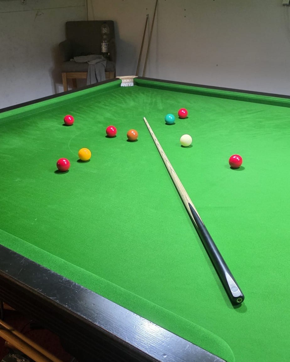 De baulkkant van een snookertafel, met de keu rustig op het laken, de kleuren tussen de rode en een stoel op de achtergrond